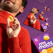 Image of man in red sweater holding an orange My/Mochi ball and laying down on purple shag carpet with a box of Sweet Mango next to him. Also in picture: a puzzle of the same image (man in red holding mochi)